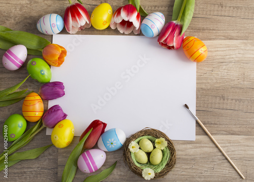 Photograph of a couple of colorful easter eggs with a bunch of tulips alligned around a sheet of white paper with copy-space on wooden surface