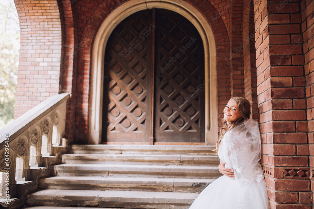 Beautiful young blond bride with bridal bouquet stands over ancient arch