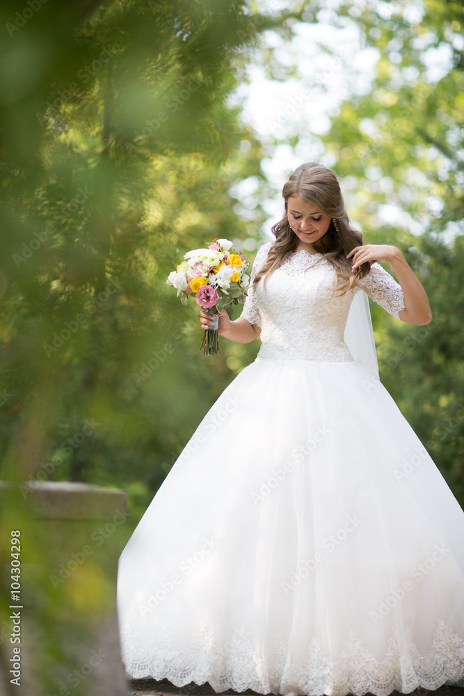 Gorgeous blonde bride in elegant dress holding bouquet and posing in the sunny summer park or garden on their wedding day Gorgeous blonde bride in elegant dress holding bouquet and posing in the sunny summer park or garden on their wedding day