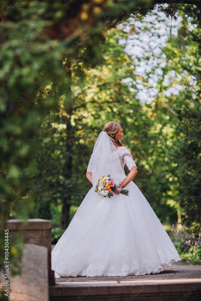 Gorgeous blonde bride in elegant dress holding bouquet and posing in the sunny summer park or garden on their wedding day