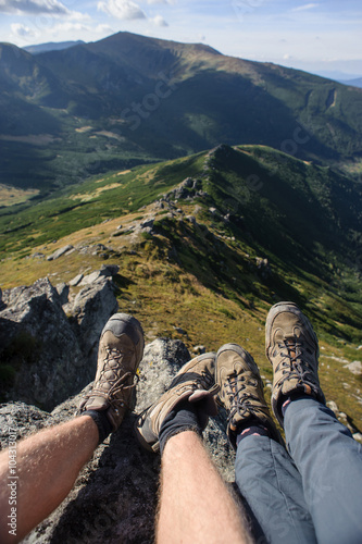 Couple's legs together on the rock on top of the mountain by the cliffside with mountain ridge on the background. Hikers boots. Summer time