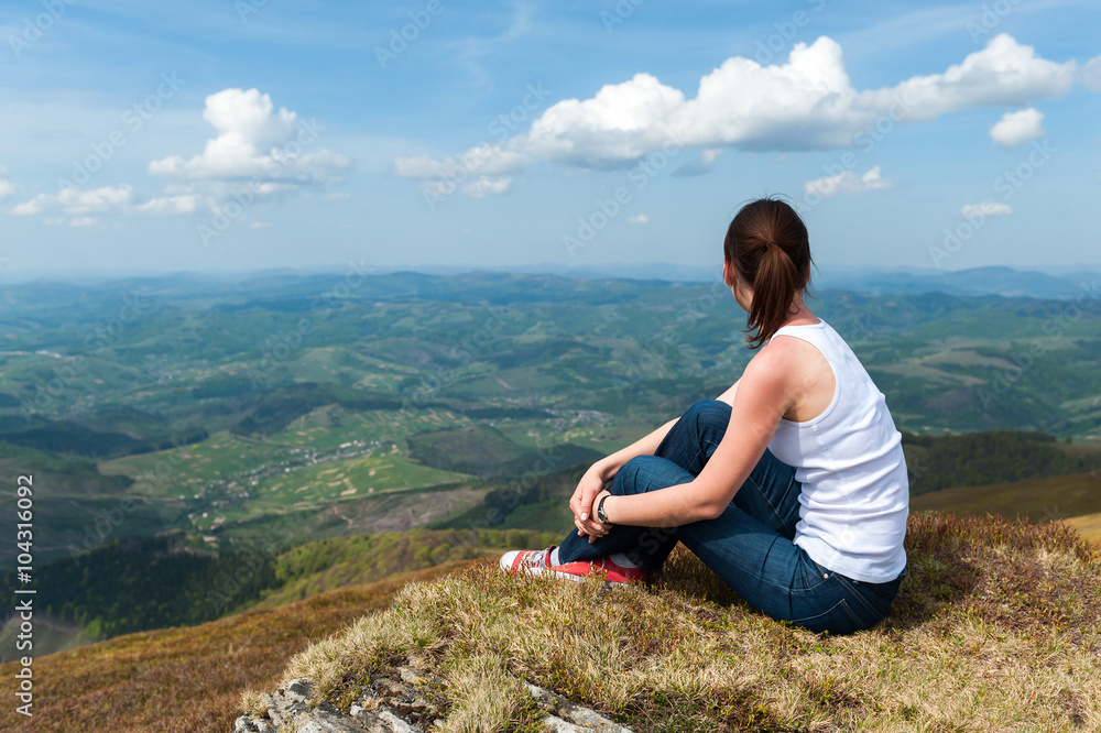 young woman sitting on top of a mountain and looking at the land
