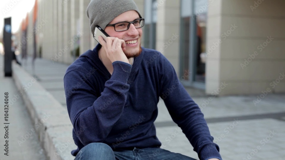 Young, handsome man in grey hat and glasses sitting near building and talking thru the phone. He has good humour. Positive emotions Young, handsome man in grey hat and glasses sitting near building and talking thru the phone. He has good humour. Positive emotions