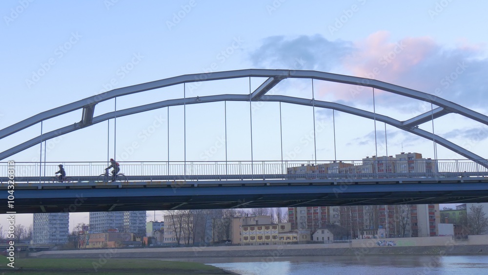 Dad and Kid Silhouettes Are Riding a Bicycles Cars Are Driving by an Arch Bridge Upon a River People on a Bridge Multi-Storeyed Houses Cityscape Evening Dad and Kid Silhouettes Are Riding a Bicycles Cars Are Driving by an Arch Bridge Upon a River People on a Bridge Multi-Storeyed Houses Cityscape Evening