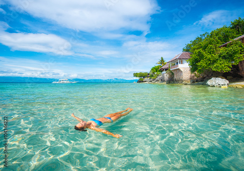 Woman in bikini relaxing lying on water against background of beach and bungalow.