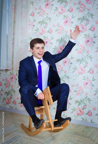 Happy childish young man in business suit riding toy wooden horse. Adult acting like a child concept