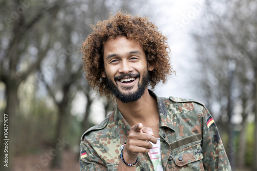 Portrait of smiling man with dyed ringlets wearing camouflage shirt pointing on viewer