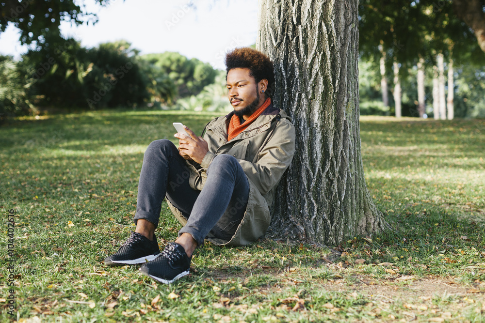 Young man with smartphone leaning against tree trunk on a meadow
