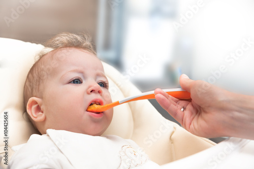 Mother feeding her happy cute little baby from spoon with light background and copy space. Child  sitting in baby seat with open mouth