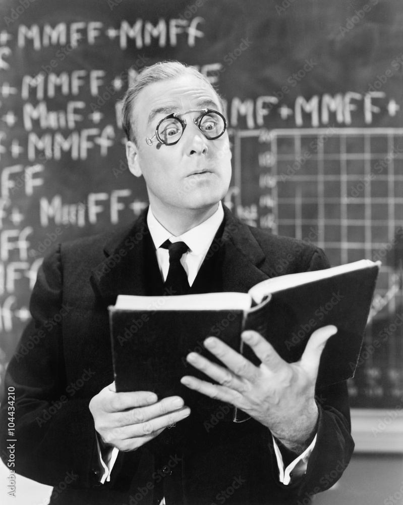 Teacher holding a book in front of a black board looking surprised Teacher holding a book in front of a black board looking surprised