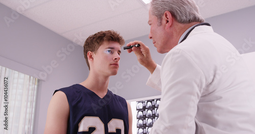 Fototapeta Senior medical doctor checking basketball player's eyes with fla