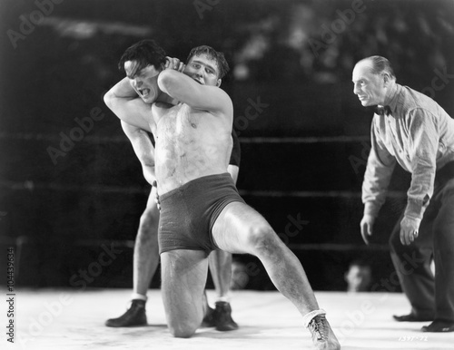 Fotografie Two men wrestling with referee making a call in the background