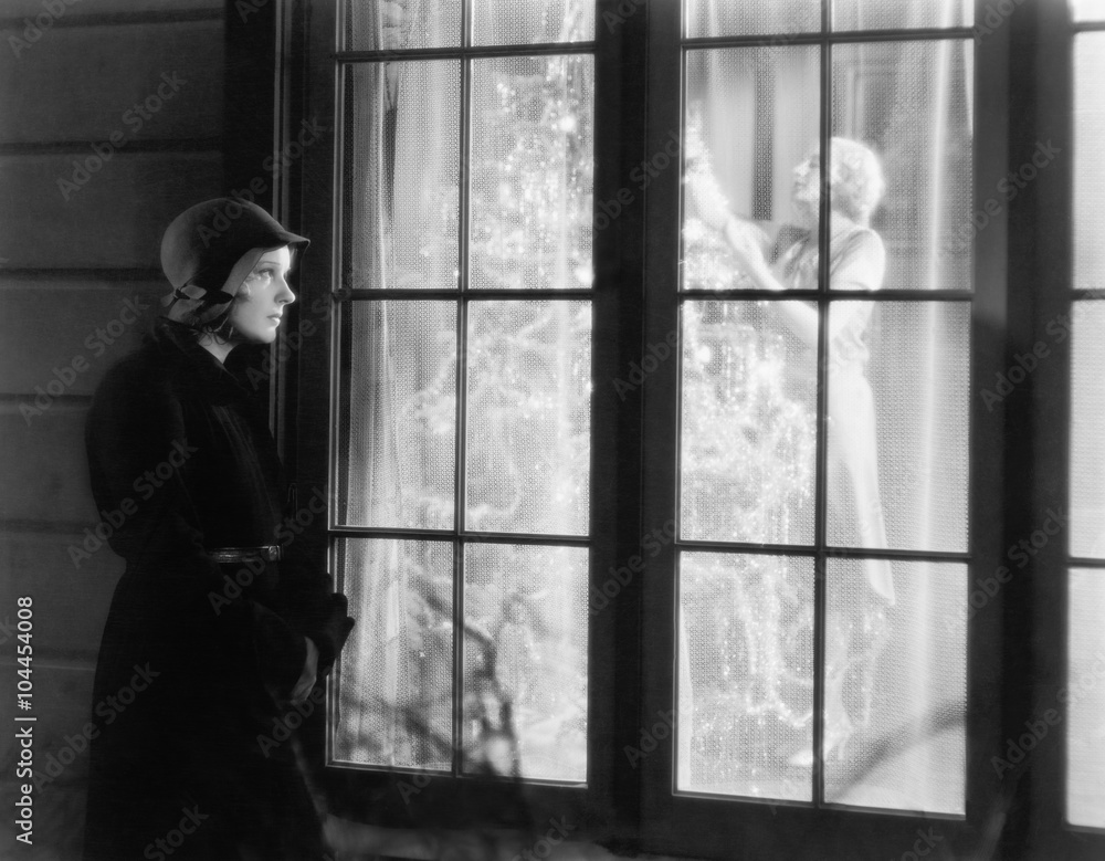 Woman standing outside of a window watching a woman trimming a Christmas tree 