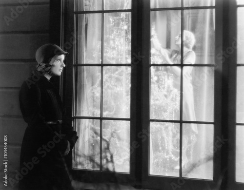 Woman standing outside of a window watching a woman trimming a Christmas tree 