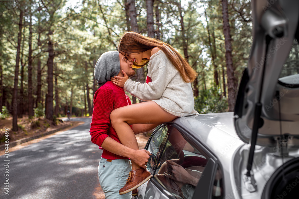 Young and lovely couple in sweates and hats having fun hugging together near the car on the roadside in the pine forest Young and lovely couple in sweates and hats having fun hugging together near the car on the roadside in the pine forest