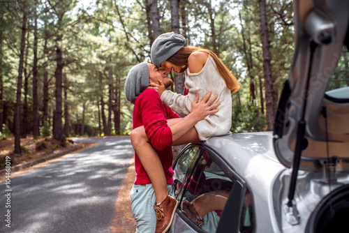 Young and lovely couple in sweates and hats having fun hugging together near the car on the roadside in the pine forest