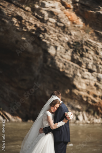 Handsome romantic groom and beautiful bride posing near river in scenic mountains