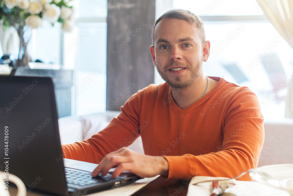 Handsome young man working on notebook, smiling, looking at camera, while enjoying coffee in cafe Handsome young man working on notebook, smiling, looking at camera, while enjoying coffee in cafe