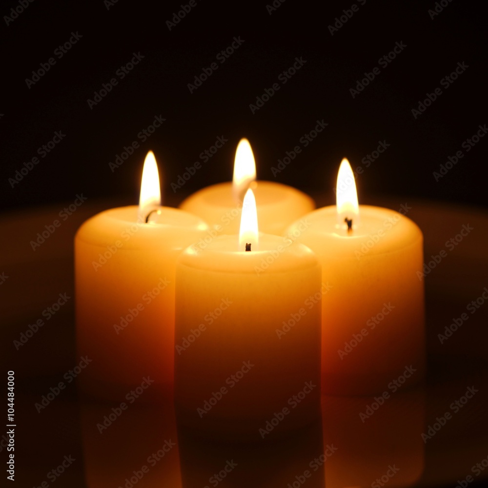 Four burning yellow candles with reflection on a white plate against dark background