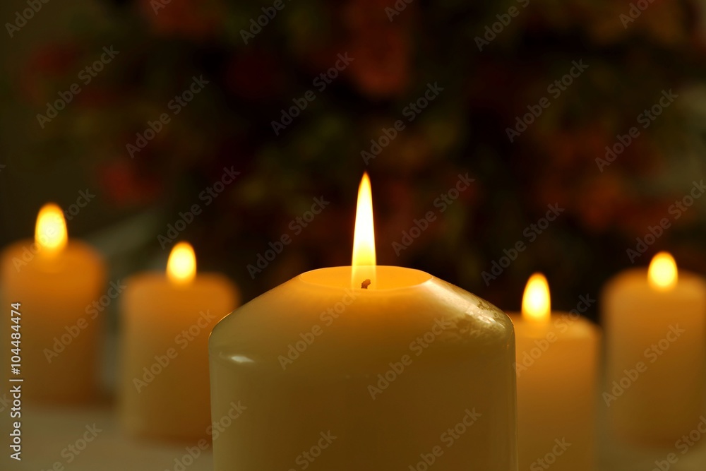 Line of burning candles with a big candle in the foreground against blurred flower background Line of burning candles with a big candle in the foreground against blurred flower background