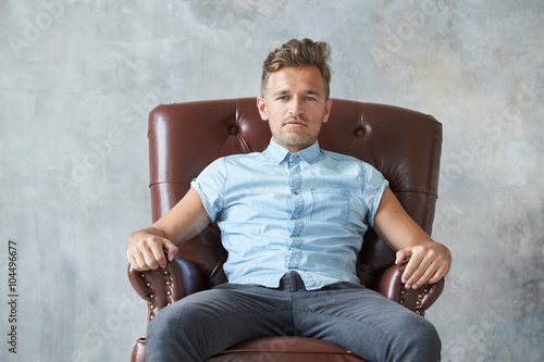 Portrait of a stylish intelligent man stares into the camera, small unshaven, charismatic, blue shirt, sitting on a brown leather chair, dialog, negotiation, short sleeve, brutal, hairstyle