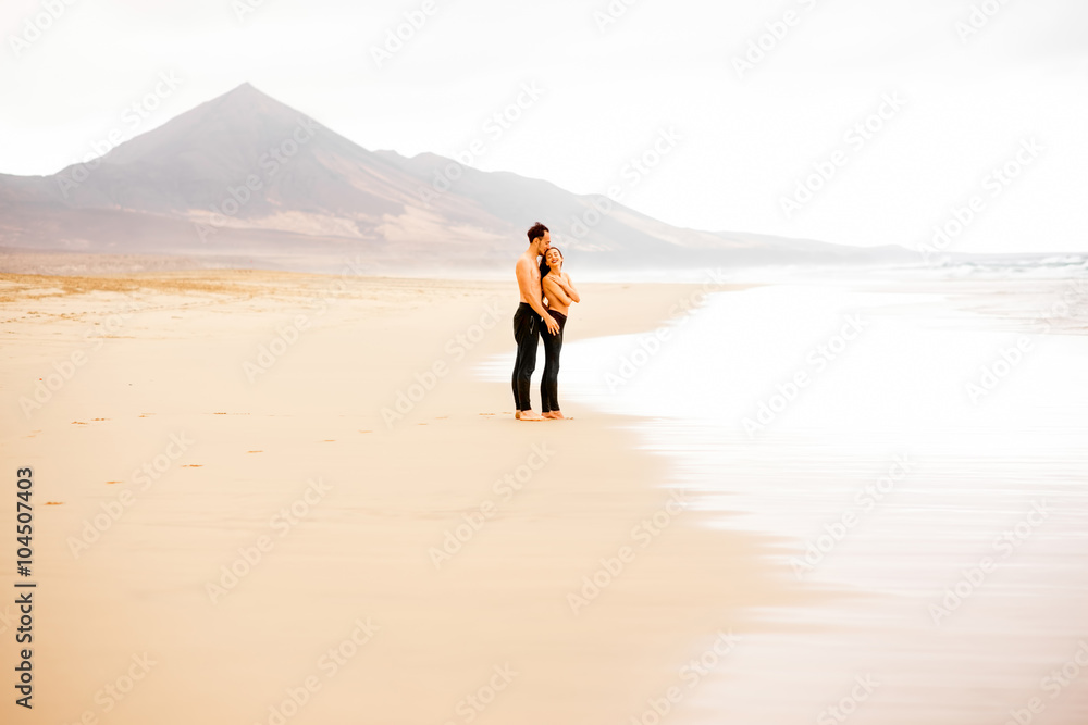 Young couple with naked torso embracing on the beautiful sandy beach with mountains on background on Fuerteventura island in Spain. General plan with copy space