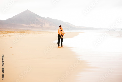 Young couple with naked torso embracing on the beautiful sandy beach with mountains on background on Fuerteventura island in Spain. General plan with copy space