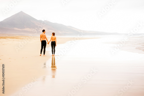 Young couple with naked torso standing and holding hands on the beautiful sandy beach with mountains on background on Fuerteventura island in Spain. General plan  rear view with copy space