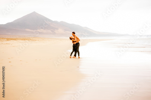 Young couple with naked torso embracing on the beautiful sandy beach with mountains on background on Fuerteventura island in Spain. General plan with copy space