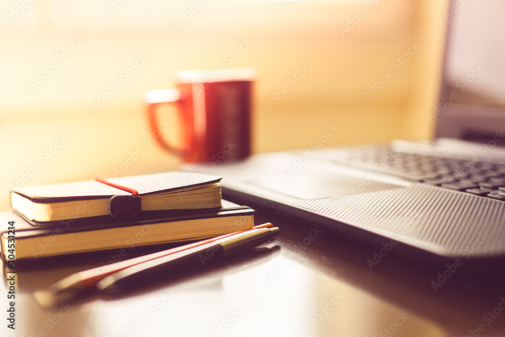 Fototapeta premium Notepads and pens with cup of coffee and laptop in background