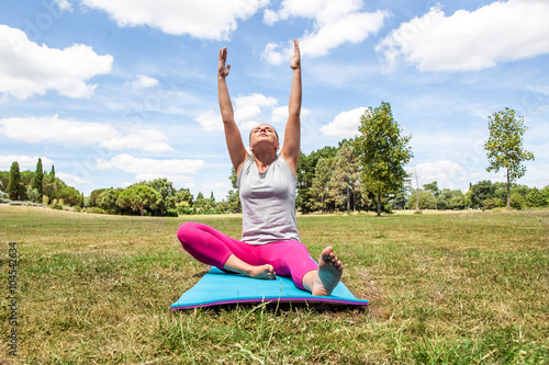 wellbeing and stretching outside - relaxed young blond woman doing yoga with arms raised on exercise mat over summer blue sky.