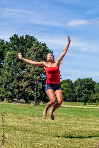 energy and fun outdoors - cheerful young blond woman laughing and jumping for success and happiness in green park, sunny summertime