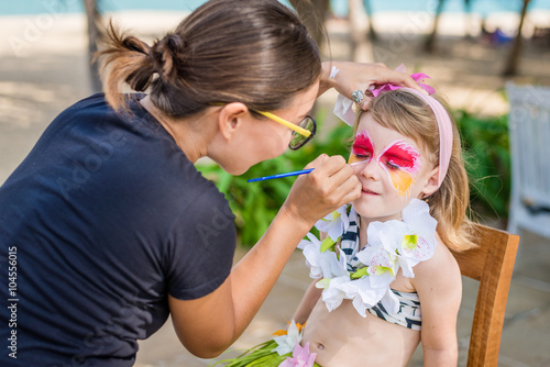 Young woman makes the child's vivid face painting. Fancy dress party on the beach.