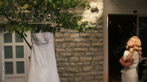 Charming blond bride in white robe smiling with eyes closed with wedding dress hanging on a tree on background photo