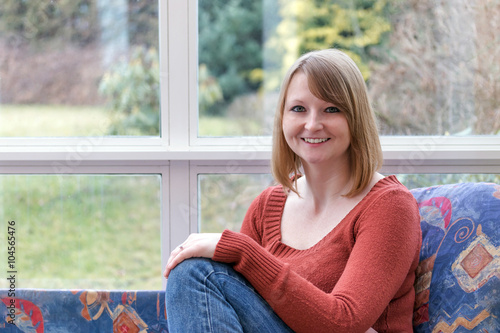 Smiling young woman dressed in a brown sweater is sitting on the sofa in conservatory and she is looking at the camera. Free Download Smiling young woman dressed in a brown sweater is sitting on the sofa in conservatory and she is looking at the camera. - GFXLoad