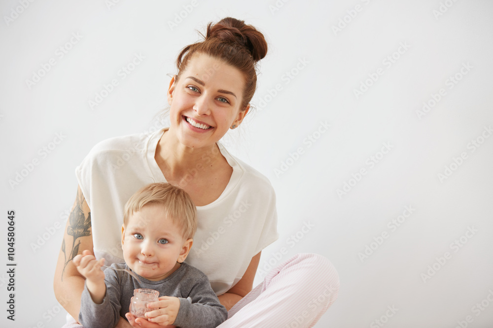 Young attractive mother with her one years old little son dressed in pajamas. Boy eating a fruit smoothie himself in the bedroom at the weekend together, warm and cozy scene. Selective focus. Young attractive mother with her one years old little son dressed in pajamas. Boy eating a fruit smoothie himself in the bedroom at the weekend together, warm and cozy scene. Selective focus.