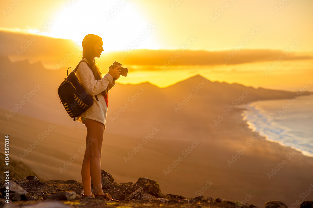 Obraz premium Young female traveler with backpack and photo camera photographing beautiful Cofete coastline with mountains on the sunset on Fuerteventura island in Spain.