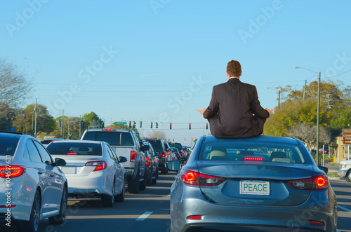 A man is dealing with intense work rush hour traffic jam stress by getting relief doing yoga on top of his car in this humorous scene that shows PEACE on the license plate of the car he is sitting on.