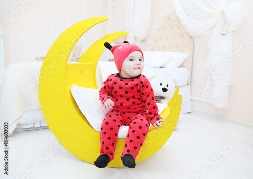 Маленький ребенок, одетый в красный костюм с черным в горошек. Детский интерьер. Белая светлая комната с кроваткой, луна игрушка, плюшевый мишка.