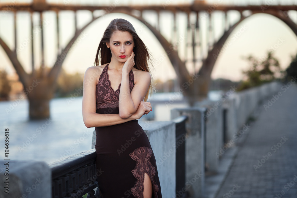 Beautiful young fashionable woman posing in dress at the river coast in the evening after sunset. Vogue style. Urban background