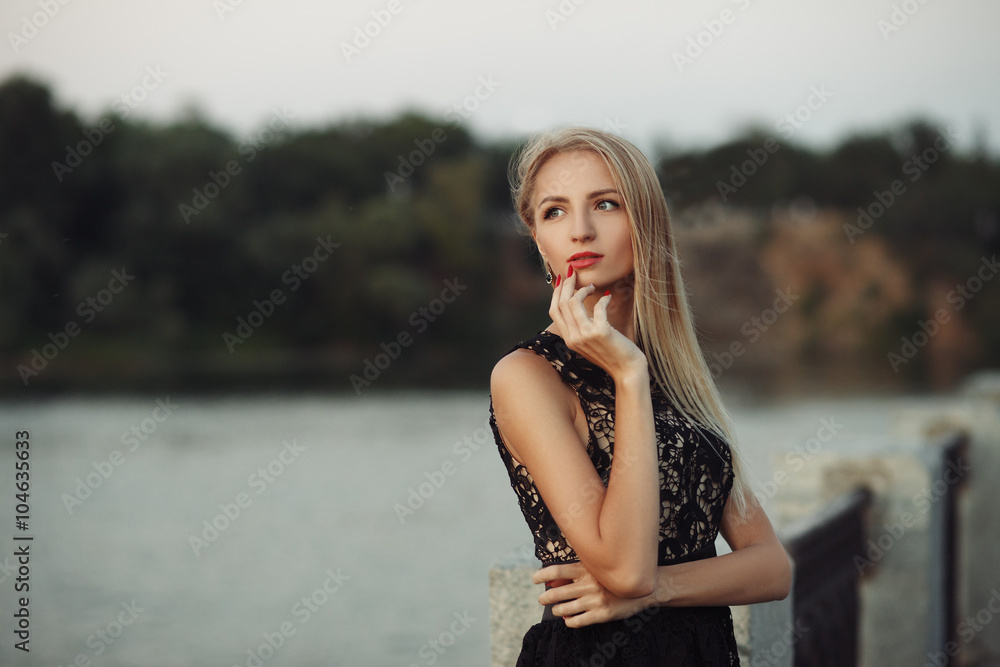 Naklejka premium Beautiful young fashionable woman posing in dress at the river coast in the evening after sunset. Vogue style. Urban background