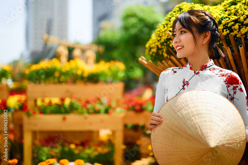 Culture Of Asia. Beautiful Happy Smiling Young Asian Woman Wearing Traditional White Ao Dai Dress ( Clothing ), Vietnamese Conical Hat ( Non La ) Leaf Hat In Flower Garden In Ho Chi Minh City, Vietnam