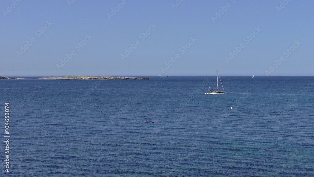 Small empty island in the sea near horizon. There is a yacht with no sail anchored between coast and island and a man in an inflatable boat by the yacht. Small empty island in the sea near horizon. There is a yacht with no sail anchored between coast and island and a man in an inflatable boat by the yacht.
