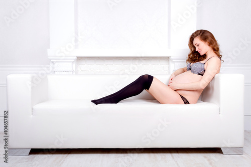 Beautiful young pregnant brunette woman / teenager in underwear is sitting on white sofa and hugs her belly