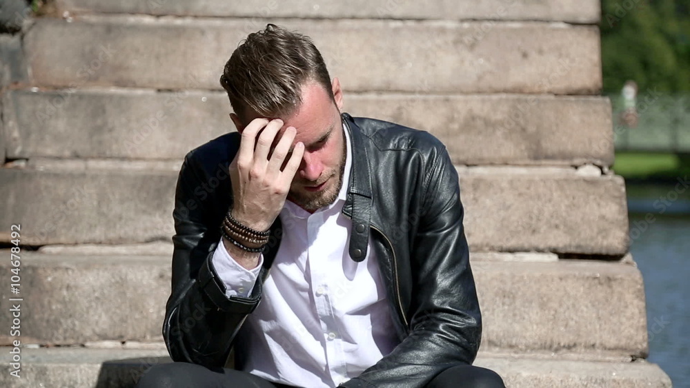 Trendy looking man sitting alone outside on steps alongside a river, wearing a black leather jacket and a white shirt. Looking away from camera with a sad look. Trendy looking man sitting alone outside on steps alongside a river, wearing a black leather jacket and a white shirt. Looking away from camera with a sad look.
