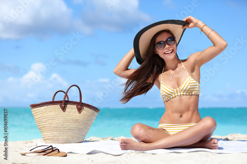 Happy beach vacation woman enjoying the sun in bikini and sunglasses having fun on a windy day holding on hat. Sexy slim Asian young adult on towel for tropical travel holidays or diet concept. photo