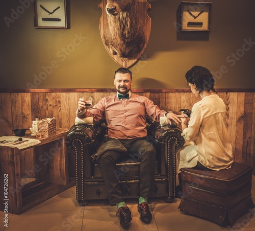 Confident old-fashioned man with glass of whisky doing male manicure in a barber shop.