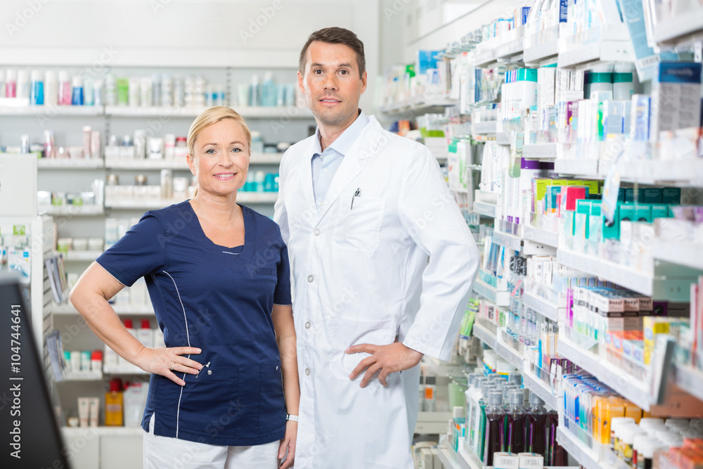Confident Pharmacist And Assistant Standing With Hands On Hip