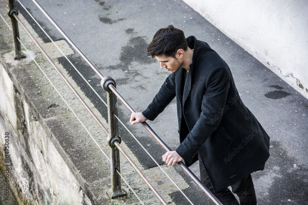 Young man in black coat looking down while leaning on railing Young man in black coat looking down while leaning on railing