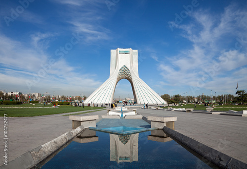 Azadi monument and its reflection on waterways in Azadi square of Tehran, against blue sky and white clouds.